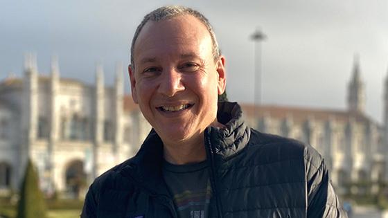 Sean Spuehler stands outdoors in front of a large building. He is smiling and wearing a black jacket and a blue t-shirt.